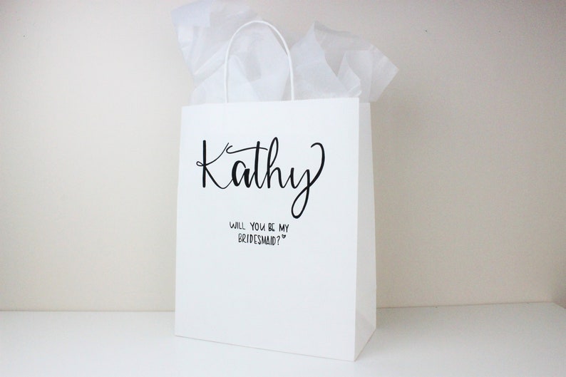 May include: White gift bag with black text that says "Kathy" and "Will you be my bridesmaid?"