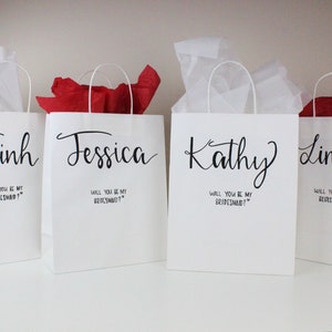 May include: Four white gift bags with black handles and text. Each bag has a name printed on the front: Trinh, Jessica, Kathy, and Linda. The text below each name reads "Will you be my bridesmaid?"