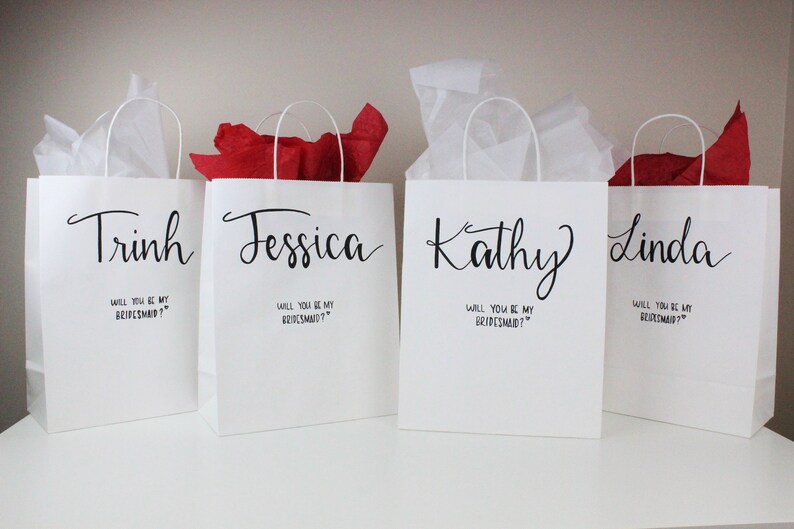 May include: Four white gift bags with black handles and text. Each bag has a name printed on the front: Trinh, Jessica, Kathy, and Linda. The text below each name reads "Will you be my bridesmaid?"