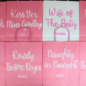 Pu&ograve; includere: Otto sacchetti regalo di carta rosa e bianchi con testi e nomi diversi su ciascuno. Il testo include "Bri's Last Bash", "Kiss Her Mass Goodbye", "Wife of the Party", "Mass to Mrs", "Bride to Be", "Rowdy Before Reyes", "Naughty in Newport" e "Bri's Bachelorette".