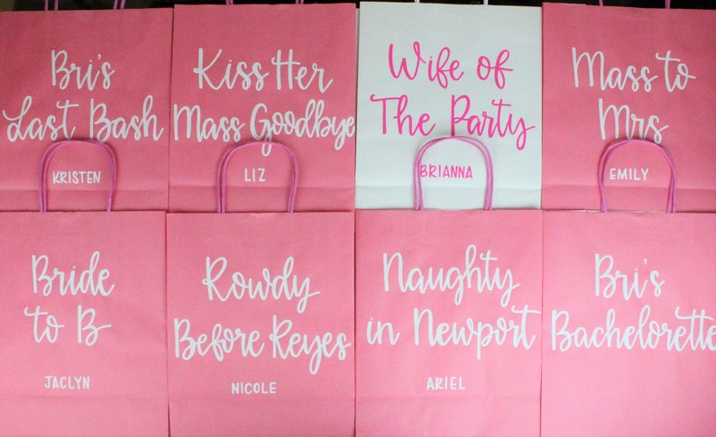 Pu&ograve; includere: Otto sacchetti regalo di carta rosa e bianchi con testi e nomi diversi su ciascuno. Il testo include "Bri's Last Bash", "Kiss Her Mass Goodbye", "Wife of the Party", "Mass to Mrs", "Bride to Be", "Rowdy Before Reyes", "Naughty in Newport" e "Bri's Bachelorette".