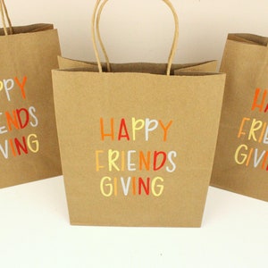 Puede incluir: Tres bolsas de regalo de papel marr&oacute;n con asas de papel retorcido. Cada bolsa tiene las palabras "HAPPY FRIENDS GIVING" impresas en letras coloridas. Las bolsas est&aacute;n dispuestas sobre una superficie blanca.