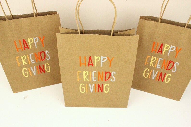Puede incluir: Tres bolsas de regalo de papel marr&oacute;n con asas de papel retorcido. Cada bolsa tiene las palabras "HAPPY FRIENDS GIVING" impresas en letras coloridas. Las bolsas est&aacute;n dispuestas sobre una superficie blanca.
