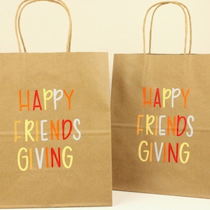 Puede incluir: Bolsas de regalo de papel kraft con las palabras "HAPPY FRIENDS GIVING" impresas en naranja, amarillo, rojo y blanco. Las bolsas tienen asas de papel trenzado marr&oacute;n.