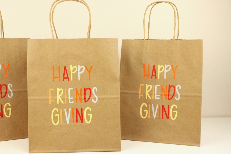 Puede incluir: Bolsas de regalo de papel kraft con las palabras "HAPPY FRIENDS GIVING" impresas en naranja, amarillo, rojo y blanco. Las bolsas tienen asas de papel trenzado marr&oacute;n.