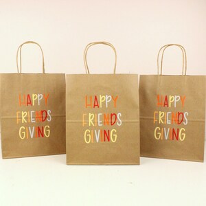 Puede incluir: Tres bolsas de regalo de papel marr&oacute;n con asas. Cada bolsa tiene las palabras "HAPPY FRIENDS GIVING" impresas en letras coloridas. Las bolsas est&aacute;n dispuestas sobre una superficie blanca.