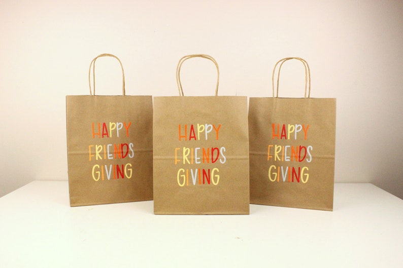 Puede incluir: Tres bolsas de regalo de papel marr&oacute;n con asas. Cada bolsa tiene las palabras "HAPPY FRIENDS GIVING" impresas en letras coloridas. Las bolsas est&aacute;n dispuestas sobre una superficie blanca.