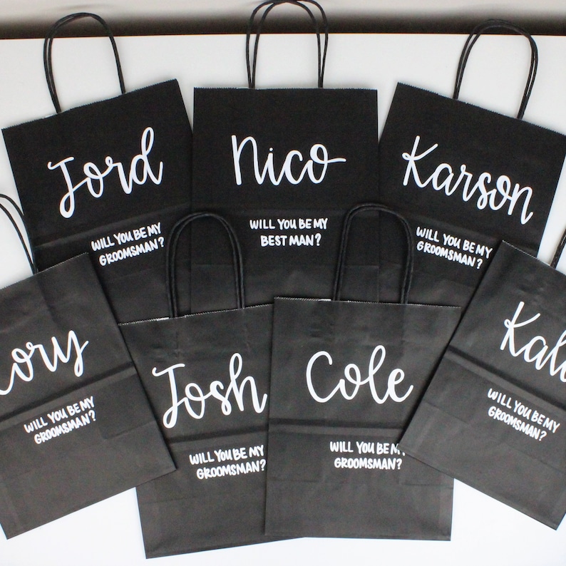 May include: Black paper gift bags with white lettering. Each bag has a name and the question "Will you be my groomsman?" or "Will you be my best man?"