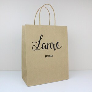 May include: A brown paper gift bag with a black handle and the name "Lanre" written in cursive with the word "Bestman" underneath.