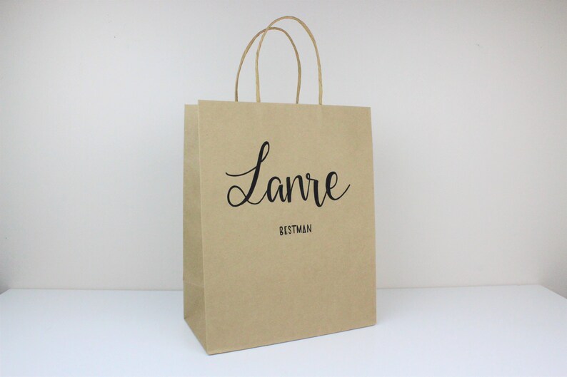 May include: A brown paper gift bag with a black handle and the name "Lanre" written in cursive with the word "Bestman" underneath.