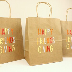 Puede incluir: Tres bolsas de regalo de papel marr&oacute;n con asas de papel retorcido. Cada bolsa tiene las palabras "HAPPY FRIENDS GIVING" impresas en letras coloridas. Las bolsas est&aacute;n sobre una superficie blanca.