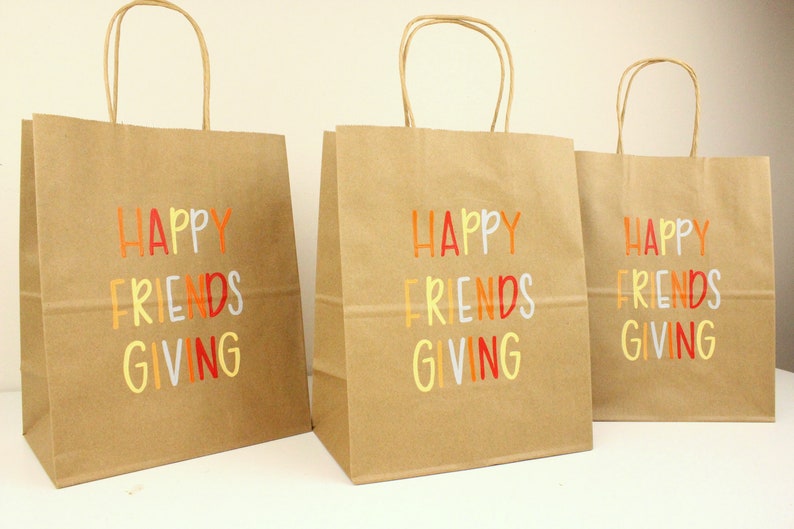 Puede incluir: Tres bolsas de regalo de papel marr&oacute;n con asas de papel retorcido. Cada bolsa tiene las palabras "HAPPY FRIENDS GIVING" impresas en letras coloridas. Las bolsas est&aacute;n sobre una superficie blanca.