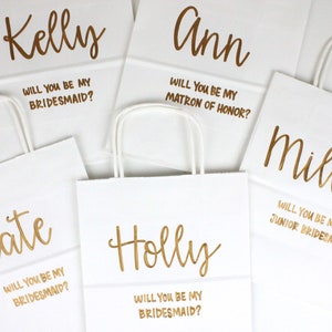 May include: Five white paper gift bags with gold lettering. Each bag has a name and a question asking the recipient to be a bridesmaid, matron of honor, or junior bridesmaid.