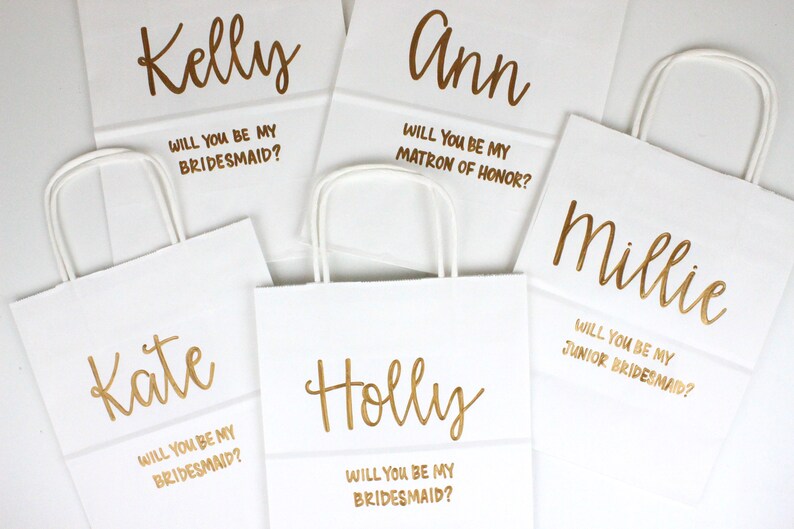 May include: Five white paper gift bags with gold lettering. Each bag has a name and a question asking the recipient to be a bridesmaid, matron of honor, or junior bridesmaid.