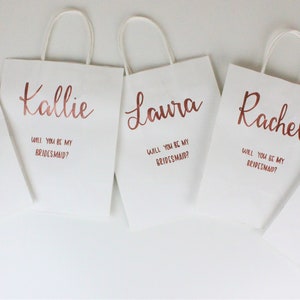 May include: Five white gift bags with rose gold lettering. Each bag has a name and the question "Will you be my bridesmaid?" printed on it.