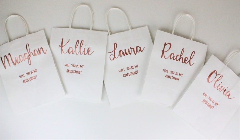 May include: Five white gift bags with rose gold lettering. Each bag has a name and the question "Will you be my bridesmaid?" printed on it.