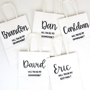 May include: Five white gift bags with black lettering. Each bag has a name printed on it, followed by the question "Will you be my groomsman?" or "Will you be my best man?"