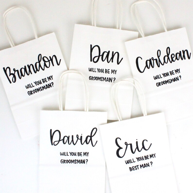 May include: Five white gift bags with black lettering. Each bag has a name printed on it, followed by the question "Will you be my groomsman?" or "Will you be my best man?"