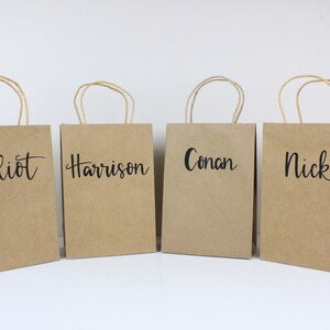 May include: Four brown paper gift bags with handles. Each bag has a name written on the front in black script: Elliot, Harrison, Conan, and Nick.