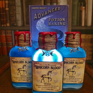 Op de afbeelding: Drie glazen flessen met blauwe vloeistof en kurkstoppen. Elke fles heeft een etiket met de tekst "The Crafty Broomstick Unicorn Blood" met een waarschuwing die luidt: "Het bloed van een eenhoorn zal je in leven houden, zelfs als je op een duim van de dood bent, maar tegen een vreselijke prijs."
