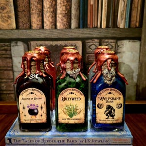 Op de afbeelding: Drie decoratieve drankflessen met etiketten met de tekst "Essence of Dittany", "Gillyweed" en "Wolfsbane Elixir". Elke fles is verzegeld met bruine was. De flessen staan op een stapel boeken.
