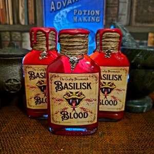Puede incluir: Tres pequeñas botellas de vidrio llenas de líquido rojo y etiquetadas con el texto "The Crafty Broomstick Basilisk Blood". Cada botella tiene un amuleto diferente colgando del cuello.