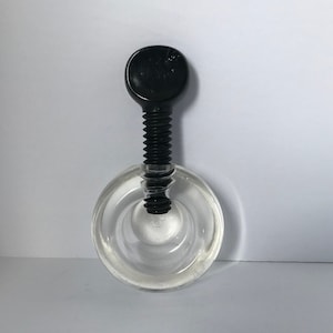 May include: A clear glass bottle stopper with a black screw top. The bottle stopper has a circular base with a raised ring around the edge.