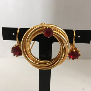 Peut inclure: Ensemble de bijoux en or avec une broche circulaire et des boucles d'oreilles assorties. La broche est composée de plusieurs anneaux d'or entrelacés avec un strass rouge au centre. Les boucles d'oreilles sont de style pendantes avec un seul strass rouge.