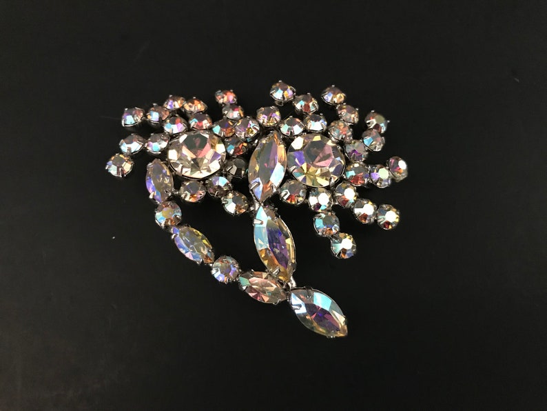 May include: A large iridescent rhinestone brooch in the shape of a flower with a long, pointed leaf extending from the bottom.