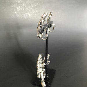 May include: A pair of silver clip-on earrings with clear rhinestones. The earrings are on a black display stand.