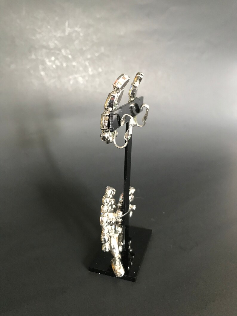 May include: A pair of silver clip-on earrings with clear rhinestones. The earrings are on a black display stand.