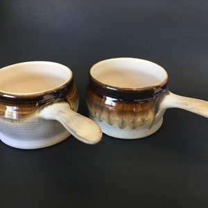 May include: Two brown and white ceramic soup bowls with handles. The bowls have a glazed finish and a rustic look.