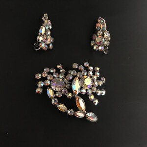 May include: A set of iridescent rhinestone jewelry featuring a brooch and a pair of earrings. The brooch is shaped like a flower with a long stem and the earrings are teardrop shaped.