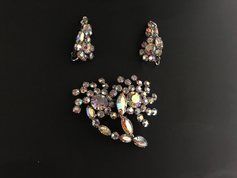 May include: A set of iridescent rhinestone jewelry featuring a brooch and a pair of earrings. The brooch is shaped like a flower with a long stem and the earrings are teardrop shaped.