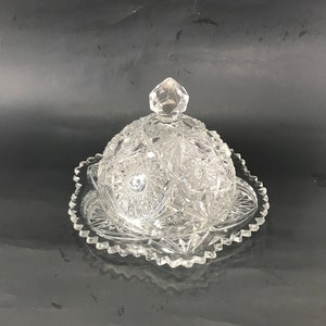 May include: A clear glass butter dish with a scalloped edge and a domed lid. The lid has a diamond-shaped knob.