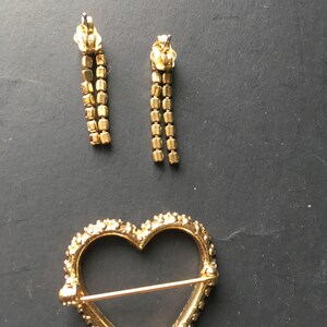 May include: A gold heart-shaped brooch with a clear glass center and a pin on the back. Two gold dangle earrings with clear stones are also pictured.