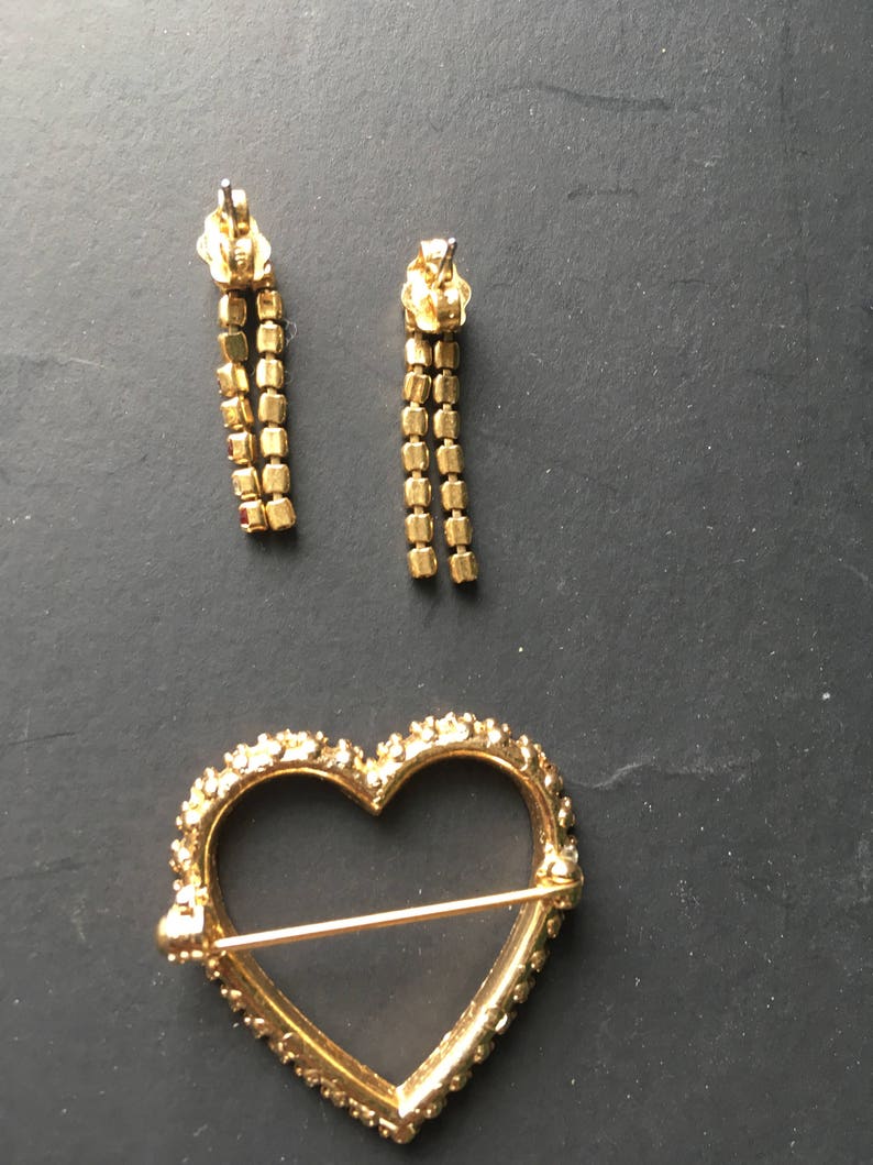 May include: A gold heart-shaped brooch with a clear glass center and a pin on the back. Two gold dangle earrings with clear stones are also pictured.