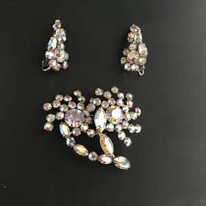 May include: A set of vintage iridescent rhinestone jewelry featuring a brooch and a pair of earrings. The brooch is shaped like a flower with a large center stone and smaller stones surrounding it. The earrings are teardrop shaped with multiple stones.