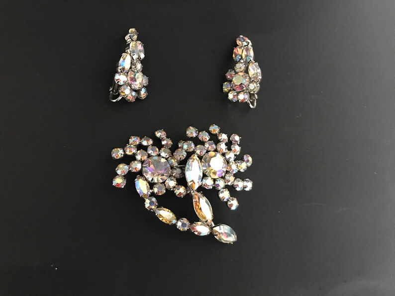 May include: A set of vintage iridescent rhinestone jewelry featuring a brooch and a pair of earrings. The brooch is shaped like a flower with a large center stone and smaller stones surrounding it. The earrings are teardrop shaped with multiple stones.