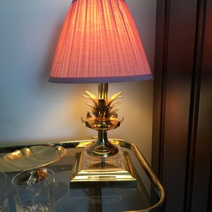 May include: A gold pineapple lamp with a pink pleated lampshade sits on a glass and gold bar cart.