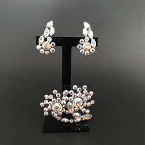 May include: A set of iridescent rhinestone jewelry featuring a brooch and a pair of earrings. The brooch is a cluster of rhinestones in a flower shape. The earrings are also flower shaped and dangle from a silver post.