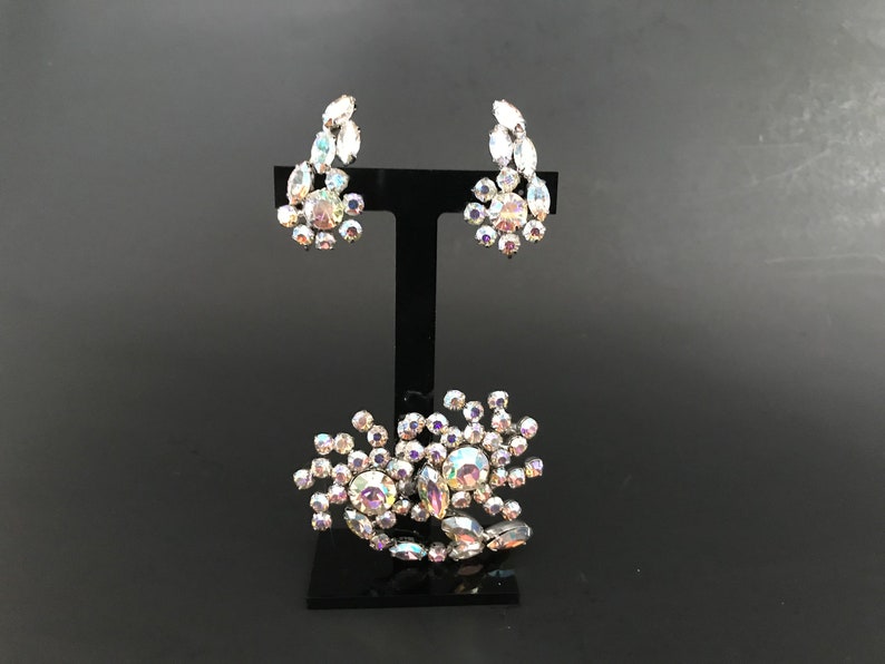 May include: A set of iridescent rhinestone jewelry featuring a brooch and a pair of earrings. The brooch is a cluster of rhinestones in a flower shape. The earrings are also flower shaped and dangle from a silver post.