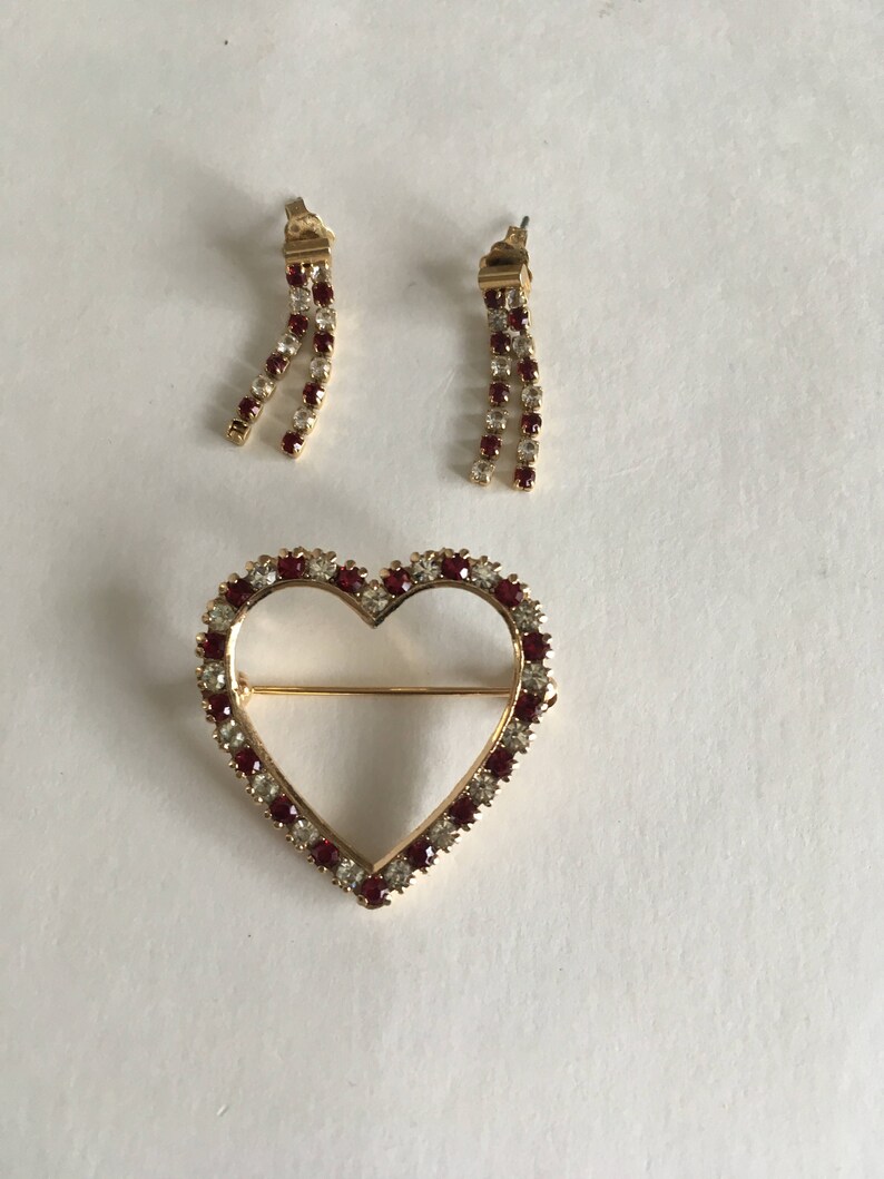 May include: A set of gold tone jewelry featuring a heart-shaped brooch and a pair of dangling earrings. The heart is outlined with clear and red rhinestones. The earrings are also adorned with clear and red rhinestones.