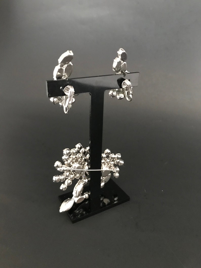 May include: A pair of silver clip-on earrings with a cluster of clear rhinestones. The earrings are displayed on a black stand.