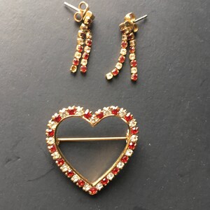May include: A gold heart-shaped brooch with red and clear rhinestones. A pair of dangle earrings with red and clear rhinestones.