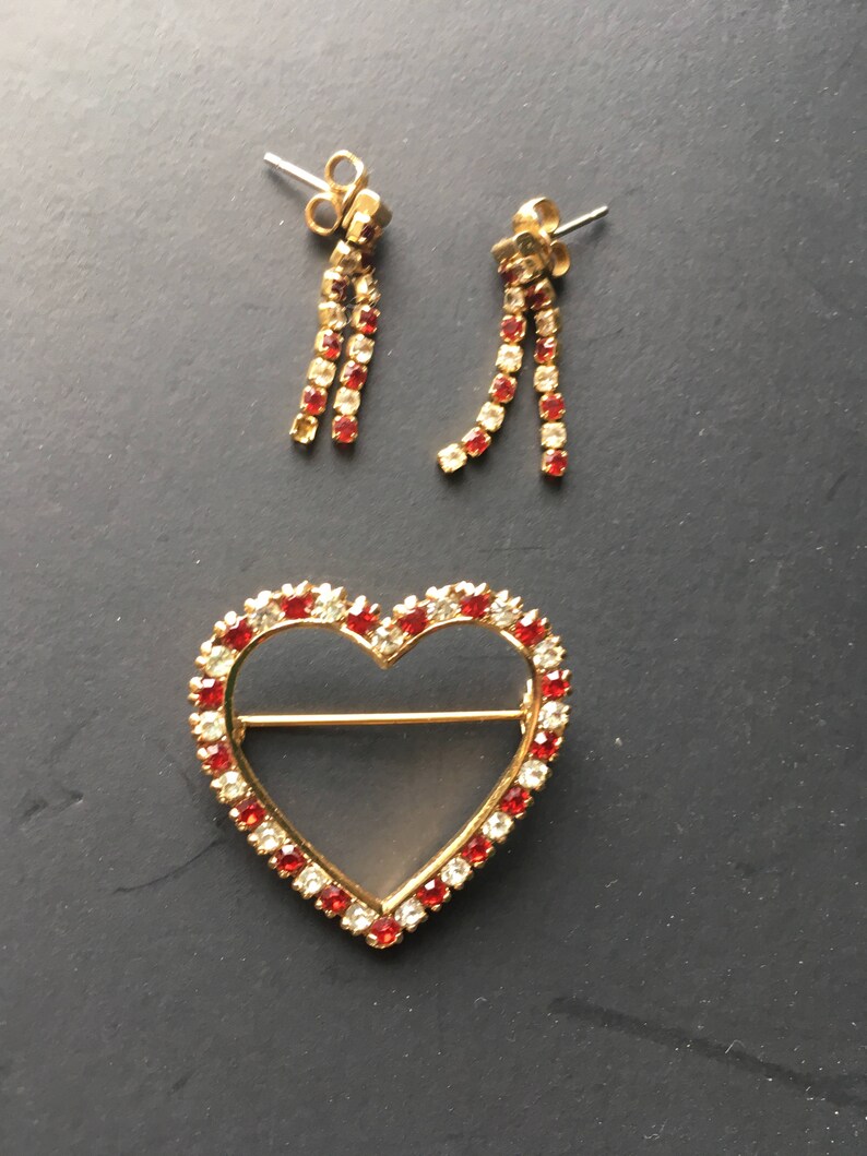 May include: A gold heart-shaped brooch with red and clear rhinestones. A pair of dangle earrings with red and clear rhinestones.