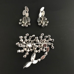 May include: A silver brooch and matching earrings with a cluster of clear rhinestones. The brooch has a pin back and the earrings have post backs.