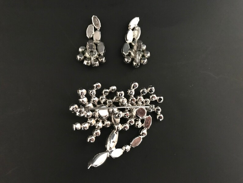 May include: A silver brooch and matching earrings with a cluster of clear rhinestones. The brooch has a pin back and the earrings have post backs.