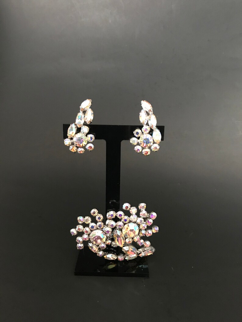 May include: A set of iridescent rhinestone earrings and a brooch. The earrings are teardrop shaped and the brooch is a cluster of rhinestones.
