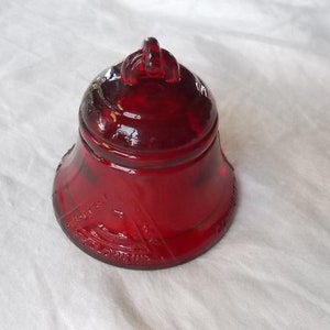 Puede incluir: Una campana de cristal rojo rubí con un asa de bucle. La campana tiene una base acampanada y una parte superior redondeada. La superficie de la campana tiene texto en relieve. La campana está sobre una superficie blanca.